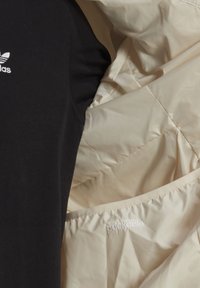 Schwarzes Longsleeve-Shirt mit weißem "adidas"-Logo, kombiniert mit einer leicht beigen Daunenjacke, die innen ein "packable"-Label hat.