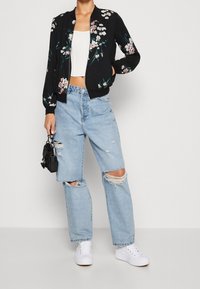 Giacca bomber nera con ricami floreali, design cropped, abbinata a jeans strappati blu chiaro e sneakers bianche. Inclusa una piccola borsa nera.