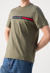 Muž v olivově zeleném tričku s krátkým rukávem značky Tommy Hilfiger s logem a červeným, bílým a námořnickým pruhem přes hrudník, spárovaném s modrými džíny.