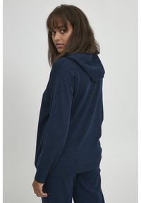 Sudadera con capucha de color azul marino, con un corte relajado, puños y dobladillo acanalados, hecha de un tejido suave y texturizado. Vista desde la parte posterior.