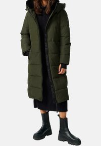 Olivengrøn polstret jakke med pelsforet hætte, lodret syning, lynlåslukning og logo-accenter. Kombineret med en sort ribstrikket kjole og sorte støvler.