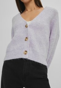 Lys lavendel cardigan med V-udskæring, ribbet tekstur, tre store brune knapper og en løs pasform, parret med sorte bukser.
