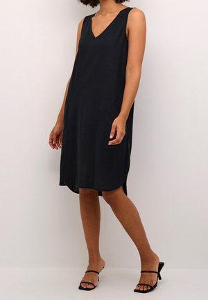 Femme portant une robe noire sans manches arrivant au genou avec un col en V et des sandales à talons noires sur un fond uni.