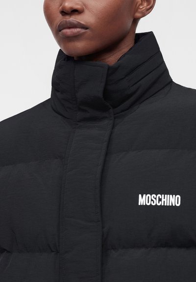 Gros plan d'une personne portant une doudoune noire à col montant avec le logo blanc Moschino sur la poitrine gauche.
