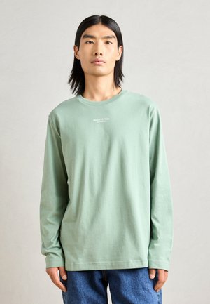 Long-sleeve, light green T-shirt made of soft cotton; features a crew neckline and small white logo on the chest. Worn with blue jeans.