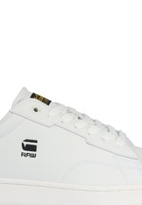 Vit sneaker med en slät lädertextur, med vita snören, kontrasterande sömmar, en svart logotyp och en gummisula med subtila detaljer.