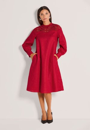 Frau steht mit Händen in den Taschen, trägt ein rotes knielanges Kleid mit Spitzenpasse und langen Puffärmeln, kombiniert mit schwarzen Absätzen.