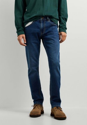 Mann trägt blaue Jeans, braune Schnürschuhe und ein grünes Langarmshirt und steht vor einem schlichten Hintergrund.