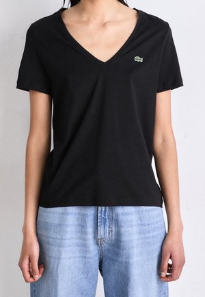Femme portant un T-shirt noir à col en V et manches courtes avec un petit logo crocodile vert, associé à un jean bleu clair taille haute.