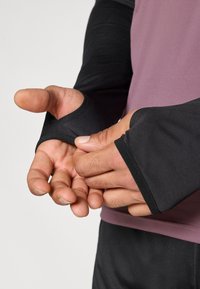 Αθλητική μπλούζα με μακριά μανίκια σε μωβ χρώμα στο σώμα και μαύρα μανίκια, με τρύπες για τους αντίχειρες στους μα νσέτες και υφή υφάσματος που είναι λεία και ελαστική.