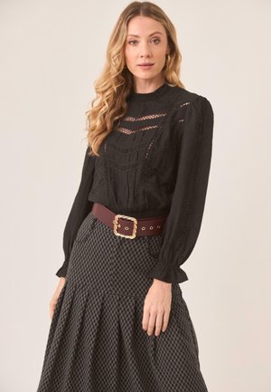 Blusa negra de manga larga con detalles de encaje, combinada con una falda gris plisada, acentuada por un cinturón ancho marrón con hebilla dorada.