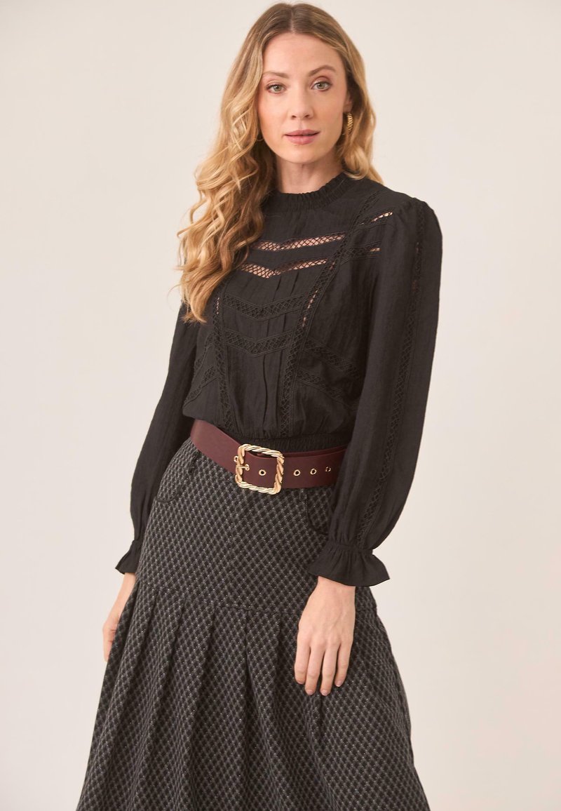 Blusa negra de manga larga con detalles de encaje, combinada con una falda gris plisada, acentuada por un cinturón ancho marrón con hebilla dorada.