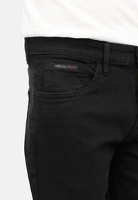 Vaqueros de mezclilla negros con un ligero brillo, que presentan un diseño tradicional de cinco bolsillos, detalles de costura y una pequeña etiqueta de marca en la cinturilla.