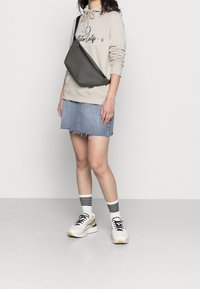 Šedá crossbody taška s elegantním designem a zipovým uzávěrem. Nositelná přes béžovou mikinu s kapucí a džínovou sukní, doplněná bílými a černými teniskami.