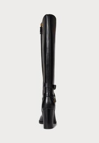 Bottes en cuir noires hauteur genou avec une finition lisse, une fermeture éclair verticale et un talon épais. Présente une sangle avec une boucle sur le côté pour le détail.