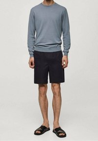 Mann in einem gedämpften blauen Langarmshirt, schwarzen knielangen Shorts und schwarzen Sandalen mit offenem Zeh, der vor einem schlichten Hintergrund steht.