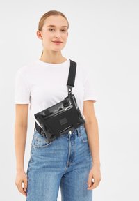 Borsa a tracolla nera e lucida con cinturino regolabile, caratterizzata da una forma rettangolare, tasca frontale e dettagli in tessuto testurizzato.