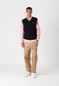 Ναυτικό γιλέκο με πλέξη καλωδίου πάνω από ένα ανοιχτό ροζ πουκάμισο με κουμπιά, συνδυασμένο με καφέ παντελόνια και λευκά αθλητικά παπούτσια. Απλός σχεδιασμός με υφασμάτινα μοτίβα.