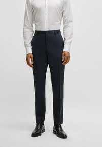 Pantaloni navy a righe con una vestibilità sartoriale, caratterizzati da una parte frontale piatta, tasche laterali e un tessuto liscio. Indossati con una camicia bianca con bottoni.