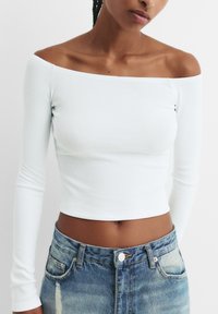 Weißes geripptes Crop-Top mit langen Ärmeln und schulterfreiem Ausschnitt. Getragen mit ausgewaschenen blauen hoch-taillierten Jeansshorts.