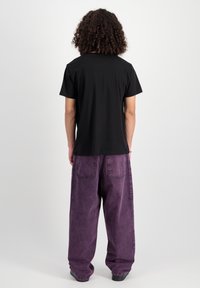Schwarzes T-Shirt mit kurzen Ärmeln, kombiniert mit locker sitzenden, lila Jeans. Der Stoff hat eine gewaschene Textur, mit sichtbaren Nähten und Taschen.