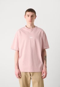 Pink cotton t-shirt with a round neckline and short sleeves. Features a small white logo patch on the chest. Loose fit and minimalistic design.