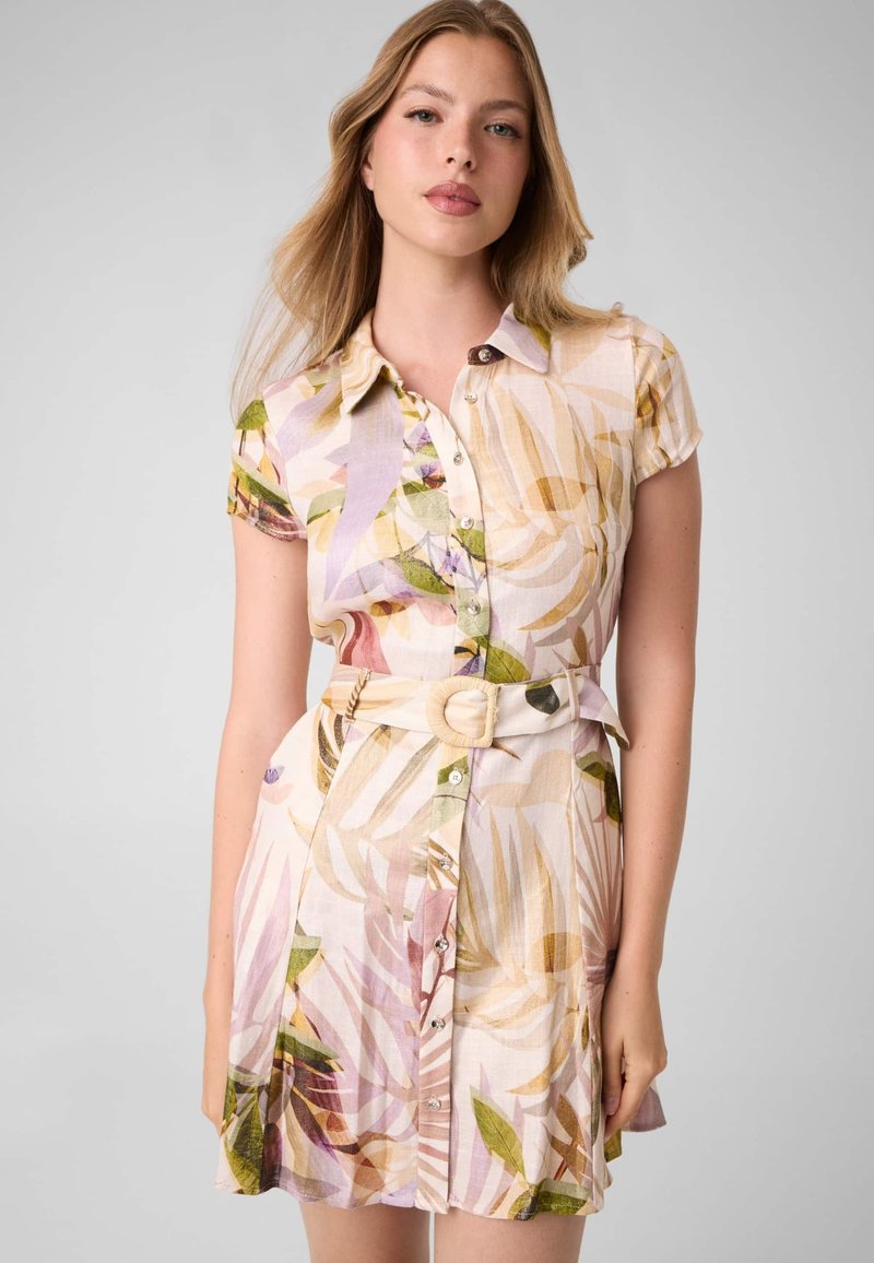 Femme aux longs cheveux châtain clair portant une robe-chemise à manches courtes et ceinture, avec un imprimé feuillage pastel, debout devant un fond uni.