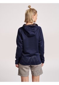 Marineblaue Kapuzenjacke mit glatter Textur und langen Ärmeln, kombiniert mit grauen Shorts mit geometrischem Muster.