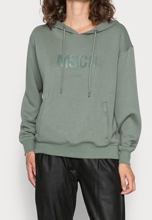 Personne portant un sweat à capuche vert discret avec des poches avant et un lettrage subtil "MSCH", associé à un pantalon noir ample.