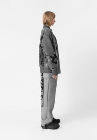 Jersey gris de punto con patrón negro, combinado con pantalones deportivos grises que tienen etiquetas laterales y zapatos negros texturizados. Fondo blanco liso.