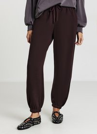 Un pantalon de survêtement bordeaux foncé avec une taille élastique et des poignets, présentant une texture lisse. Accompagné de chaussures noires ornées d'œillets en argent.