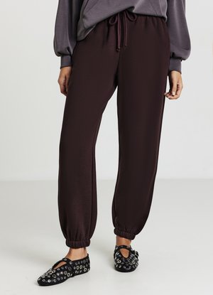 Un pantalon de survêtement bordeaux foncé avec une taille élastique et des poignets, présentant une texture lisse. Accompagné de chaussures noires ornées d'œillets en argent.