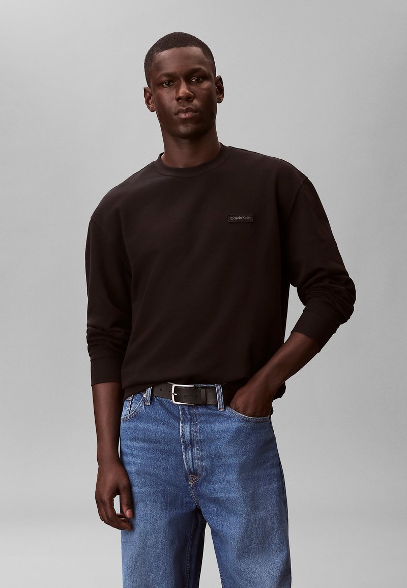 Jeune homme portant un sweat-shirt à manches longues noir Calvin Klein et un jean bleu avec une ceinture noire, debout avec une main dans la poche, sur un fond gris.