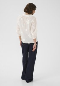Weiße Bluse mit floraler Stickerei, elastischen Ärmeln und gerafftem Ausschnitt, kombiniert mit dunkelblauen, weiten Jeanshosen.