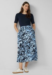 Marineblauwe fitted top met korte mouwen, gecombineerd met een blauwe maxi rok met bloemenprint, voorzien van een elastische taille en zijzakken. Witte sneakers.