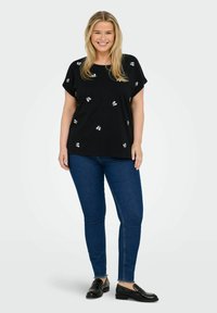 Svart kortärmad topp med vit blomsterbroderi, kombinerad med blå smala jeans och svarta loafers. Slät tyg med en avslappnad passform.