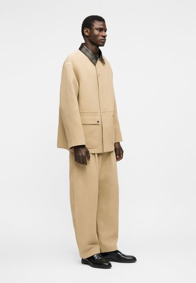 Άνδρας που στέκεται, φοράει μπεζ φαρδιά ζακέτα με μαύρο γιακά, αντίστοιχο φαρδύ παντελόνι και μαύρα παπούτσια, μπροστά σε λευκό φόντο.