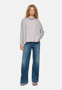 Helles graues Oversized-Sweatshirt mit einer Fronttasche, kombiniert mit weit geschnittenen blauen Jeans mit verzierten Details am Saum.