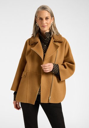 Vrouw met grijs haar draagt een camelbruine jas met een shearlingkraag en een zwart patroonhemd, staand tegen een effen witte achtergrond.