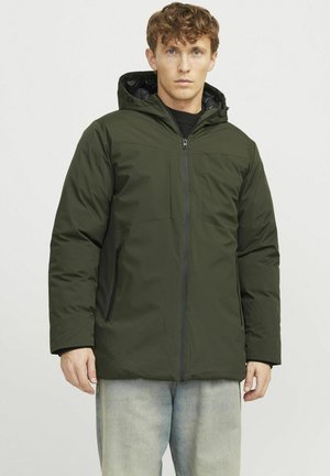 Chaqueta acolchada verde oliva con capucha, que presenta un cierre frontal, dos bolsillos laterales y una textura suave. Llevada sobre un suéter negro.
