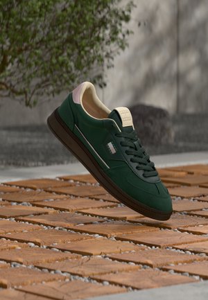 Donkergroene sneaker met zwarte veters en roze details, zwevend boven een houten betegelde vloer buiten met een wazige achtergrond.