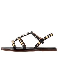 Sandalias de cuero negro con acentos de tachuelas, que presentan tachuelas metálicas en oro y plata, correa ajustable en el tobillo y suela plana marrón.