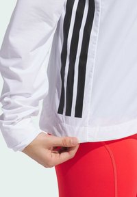 Veste de sport blanche avec des rayures verticales noires sur le côté, en tissu texturé. Une main ajuste l'ourlet près de leggings rouge vif.