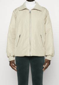 Lys beige pufferjakke med blød krave, lynlås foran og elastiske manchetter. Har to lynlåslommer på siden og en glat tekstur.