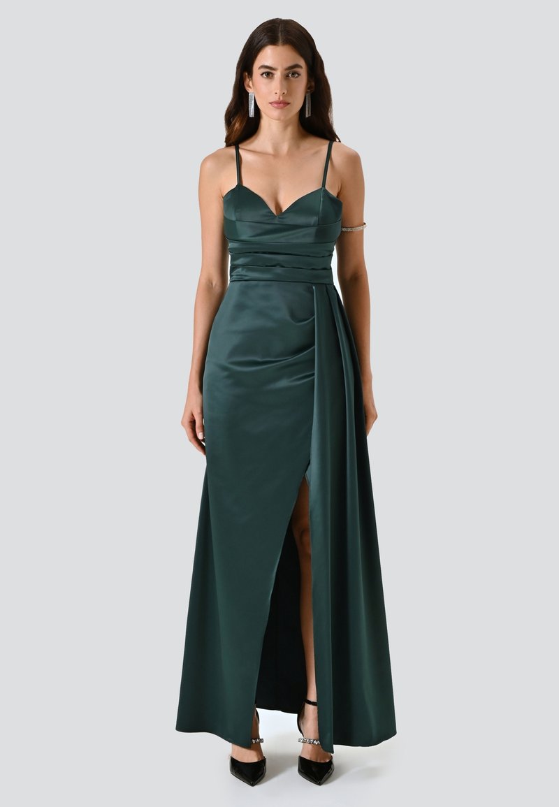 Lungo abito in satin verde smeraldo con bustino aderente, spalline sottili e uno spacco alto fino alla coscia. Presenta dettagli arricciati e una gonna fluente.