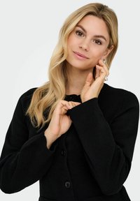 Melna kardigana, kas izgatavots no mīkstas auduma, iezīmējoties ar ribota tipa aprocēm, apaļu kakla izgriezumu un pogām, valkā cilvēks, kurš skar savu seju.