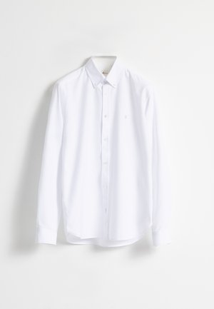 Camisa blanca de manga larga con botones y cuello de botón colgada contra un fondo blanco liso.