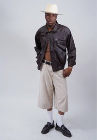 Schwarze Lederjacke, beiger weiter Shorts, weiße Socken und schwarze Schuhe. Akzentuiert mit einem Strohhut. Einfacher Hintergrund, fokussiert auf das Outfit.