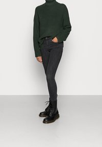 Osoba ubrana w ciemnozielony, grubo dziany sweter, czarne obcisłe dżinsy i czarne, sznurowane botki wojskowe, stojąca z jedną ręką w kieszeni.