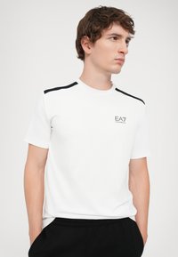 White short-sleeve t-shirt featuring black accents on shoulders and a small printed EA7 logo on the left chest. Smooth, lightweight fabric.
